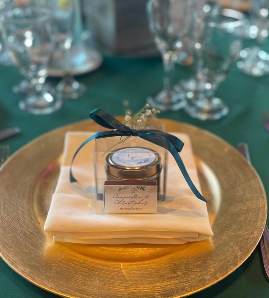 Wedding favor on top of a gold plate. Clear box with gold candle and a personalized matchbox with names. The clear box is decorated with a teal satin ribbon bow and tiny white flowers.